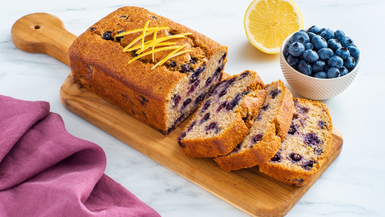 Blueberry Lemon Breakfast Loaf on a cutting board with a bowl of blueberries and a lemon on the side.
