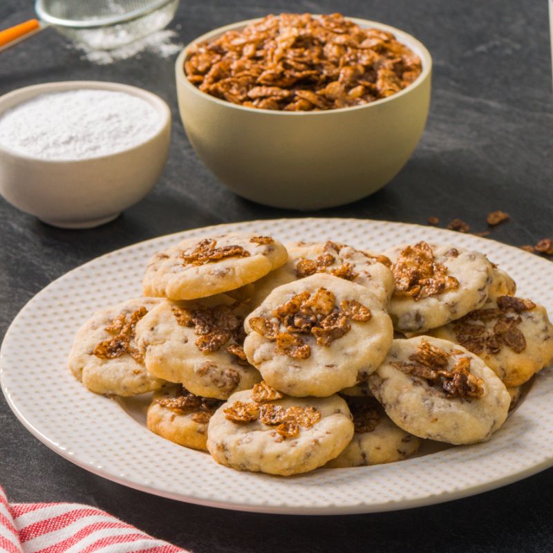 Cocoa pebbles shortbread cookies on a white plate with two bowls of ingredients in the background.