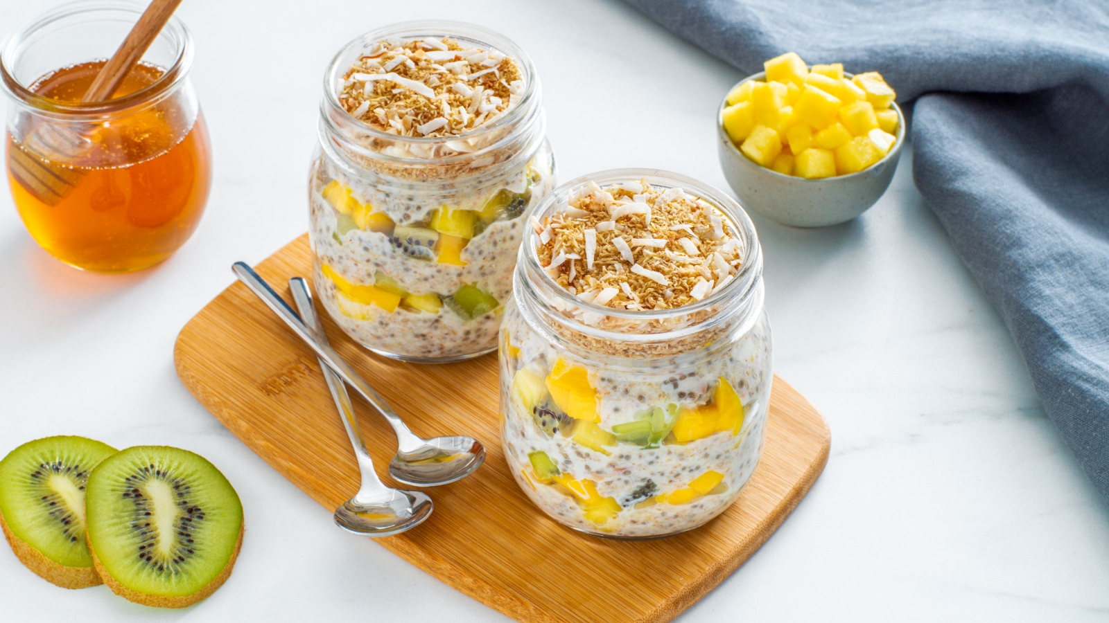 Coconut Chia and Shredded Wheat Pudding in mason jars on a wooden cutting board with kiwi slices, a jar of honey and diced fruit in the background.
