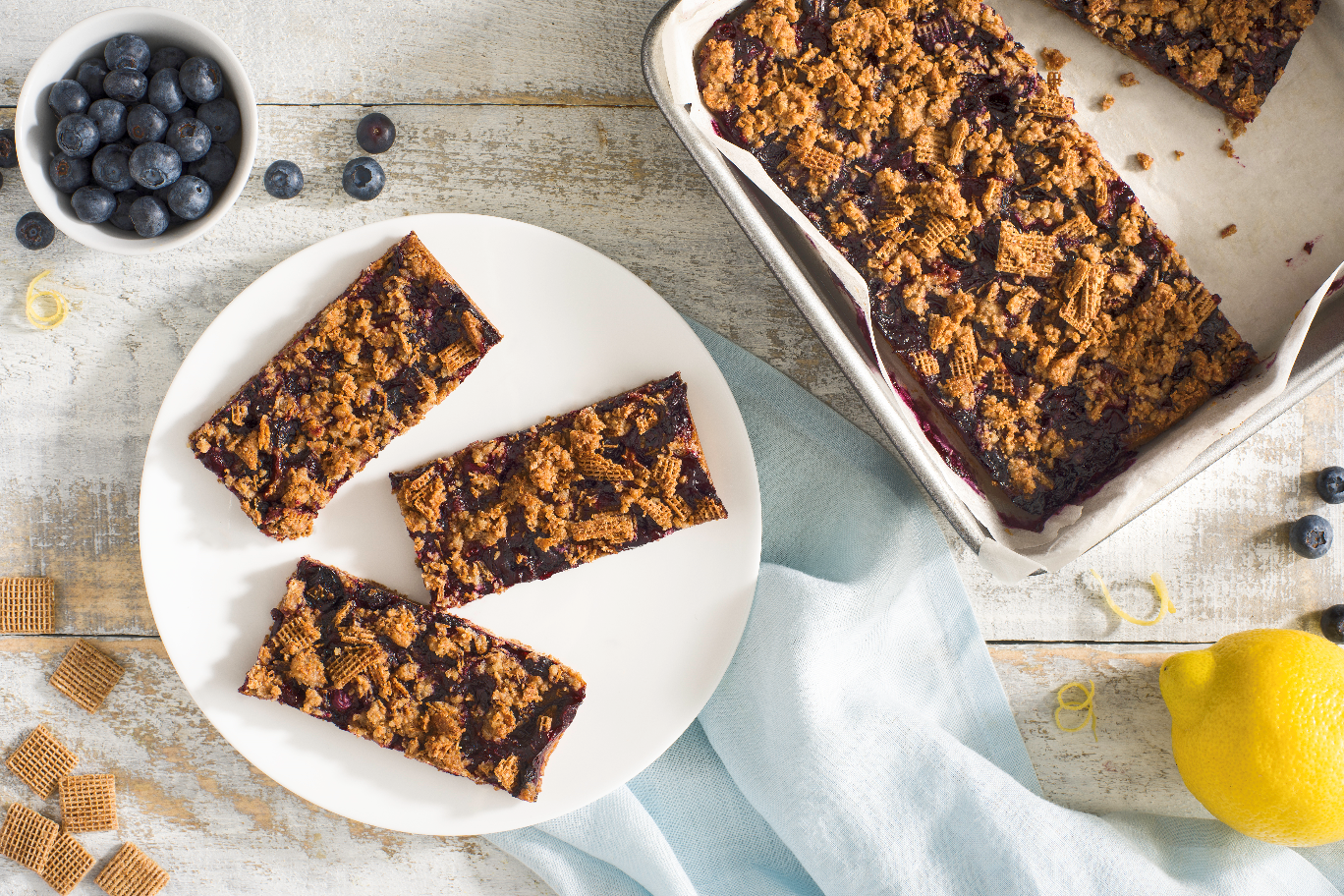 Shreddies Blueberry Crumble Bar on a white plate with a bowl of blueberries and a lemon on the side.