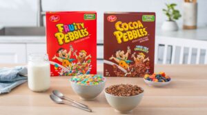 Fruity Pebbles and Cocoa Pebbles cereal boxes sitting on a kitchen table with a jar of milk, two bowls of cereal and a plate of cut up fruit.