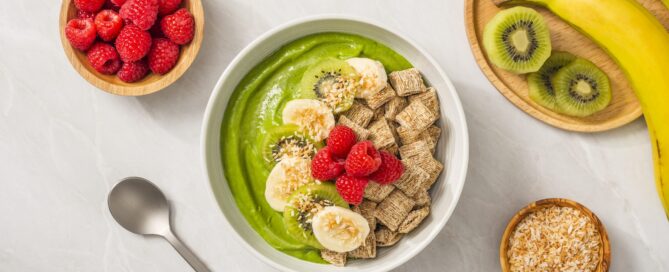 Green Goddess smoothie made with Shredded Wheat in a white bowl with bannana slices and raspberries on top. Sliced kiwi, banannas and raspberries in seperate bowls