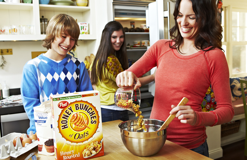 A woman and two teenagers working together in their kitchen to make a recipe with the Honey Bunches of Oats box on the table.