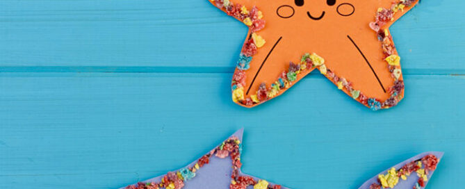 Blue paper shark and an orange paper starfish on a blue background, outlined with crushed Fruity Pebbles cereal.