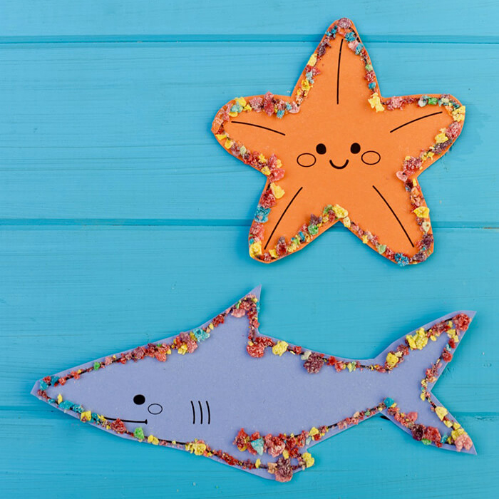 Blue paper shark and an orange paper starfish on a blue background, outlined with crushed Fruity Pebbles cereal.