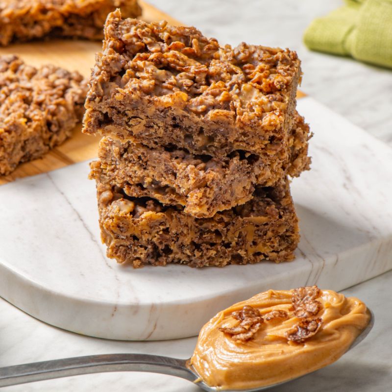 No bake Cocoa pebbles peanut butter bars sitting on a white marble cutting board with a spoon of peanut butter in the foreground.