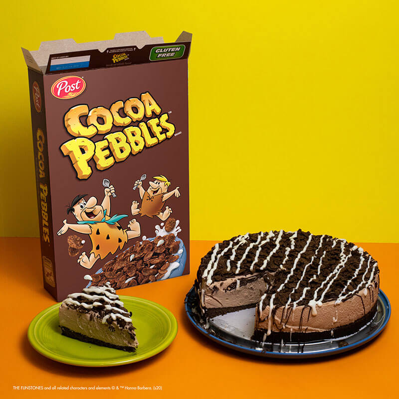 Cocoa Pebbles box and cheesecake on a yellow and orange background. 