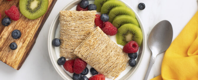 Shredded Wheat Fruitalicious in a white bowl with sliced kiwi, blueberries and raspberries.