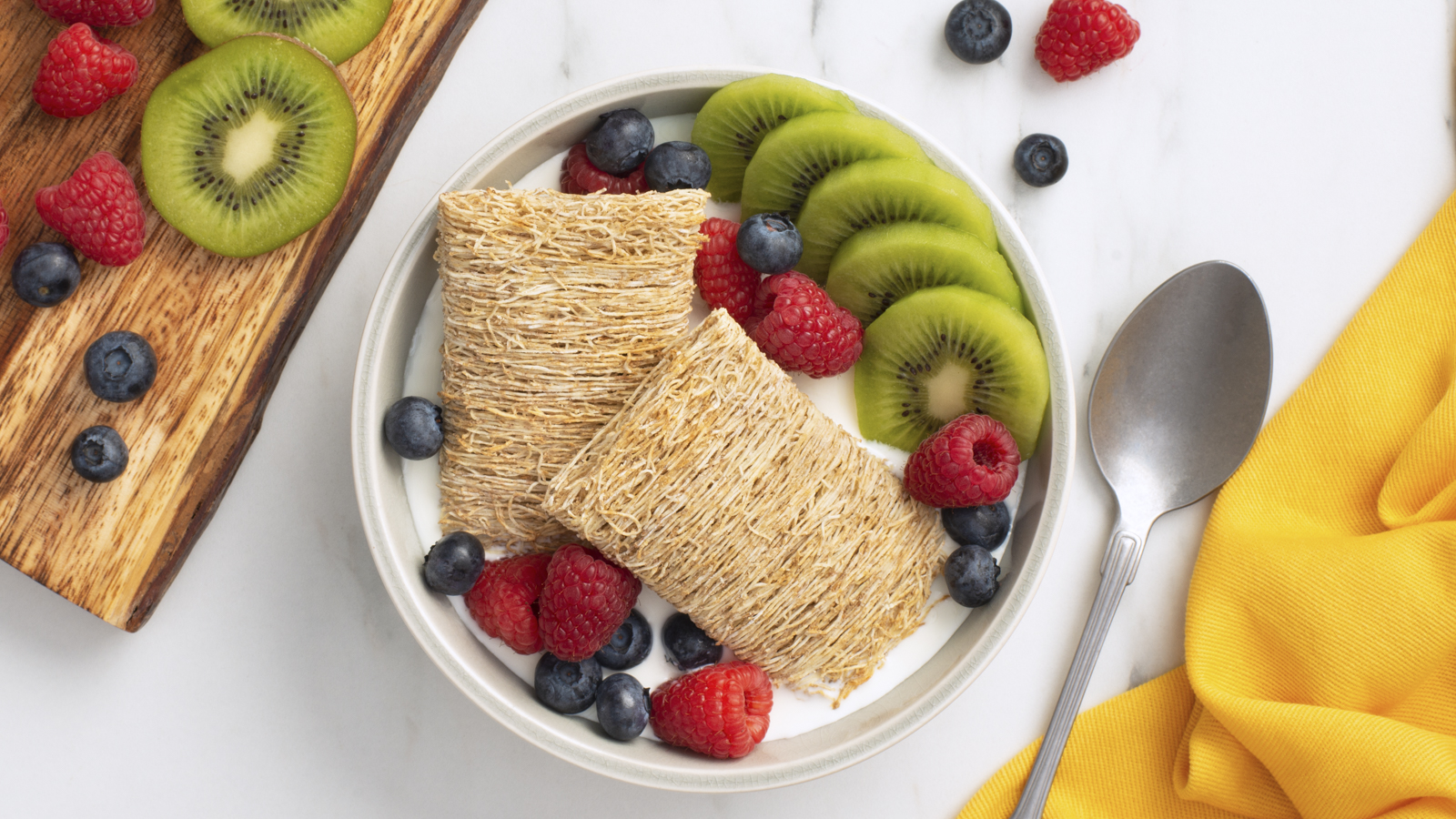 Shredded Wheat Fruitalicious in a white bowl with sliced kiwi, blueberries and raspberries. 