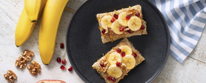 Two shredded wheat biscuits on a black plate with banana slices and pomegranate pieces.