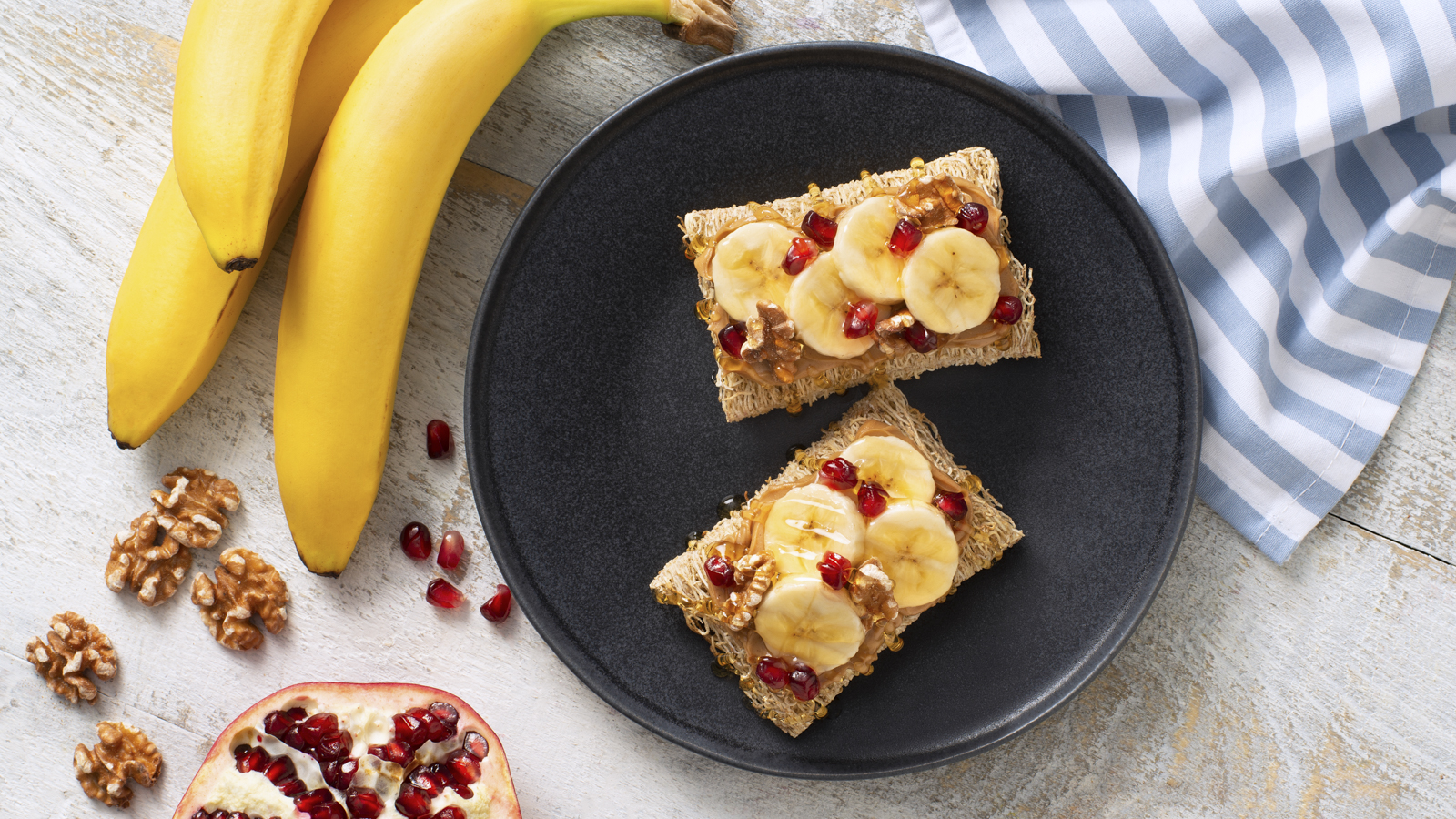Two shredded wheat biscuits on a black plate with banana slices and pomegranate pieces.