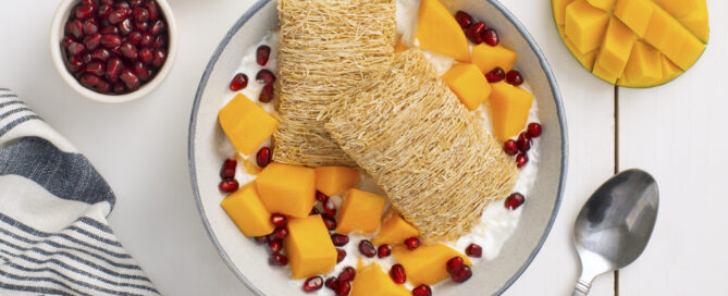 Two Shredded Wheat biscuits in a white bowl with mango pieces and pomegranate pieces.