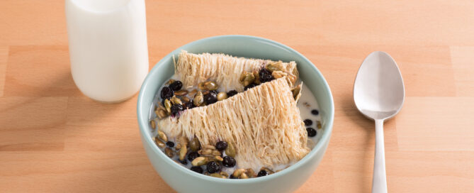 Shredded Wheat with Warm Milk, Caramelized Seeds, and Honeyed Blackcurrants in a blue bowl on a wooden table.