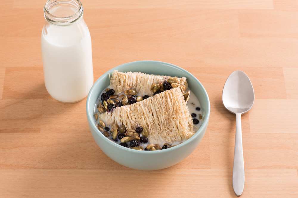 Shredded Wheat with Warm Milk, Caramelized Seeds, and Honeyed Blackcurrants in a blue bowl on a wooden table.