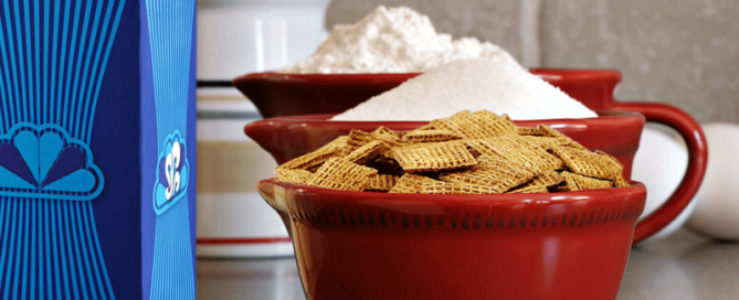 A red bowl of Shreddies sitting on a countertop next to a blue carton.