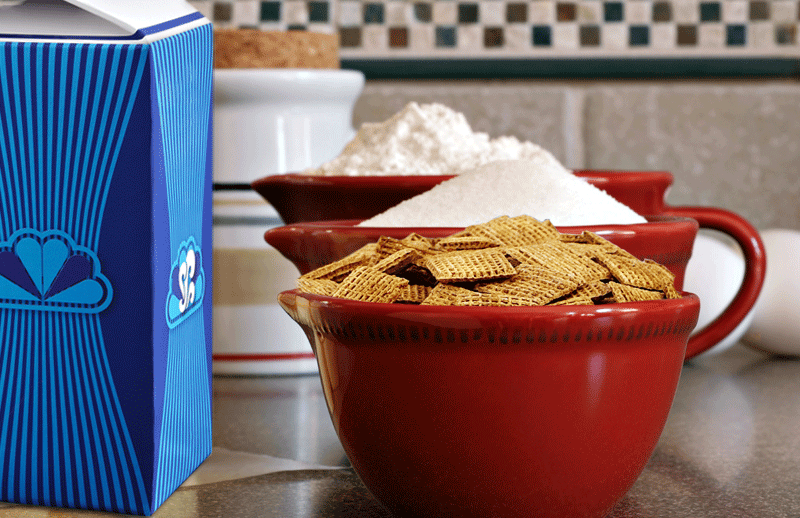 A red bowl of Shreddies sitting on a countertop next to a blue carton.