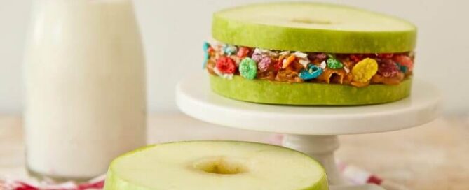 Fruity pebbles apple stackers sitting on a white platter and plate with a white and red cloth beneath them and a jug of milk in the background.