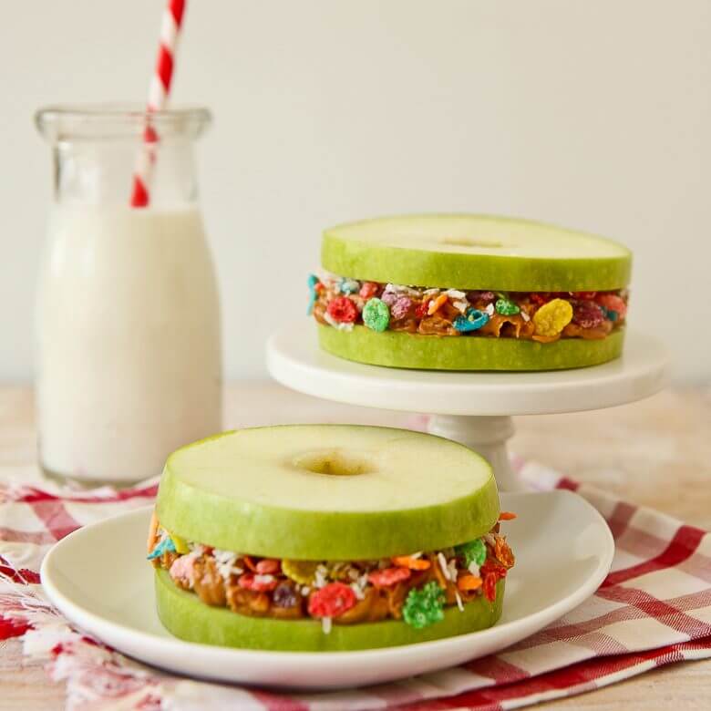 Fruity pebbles apple stackers sitting on a white platter and plate with a white and red cloth beneath them and a jug of milk in the background.