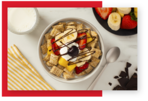 Spoon size shredded wheat in a white bowl with banana slices, strawberry slices and pineapple pieces on top.