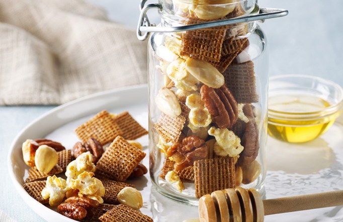 Shreddies Honey Nut Crunch Mix in a mason jar on a serving platter with a bowl of honey in the background.