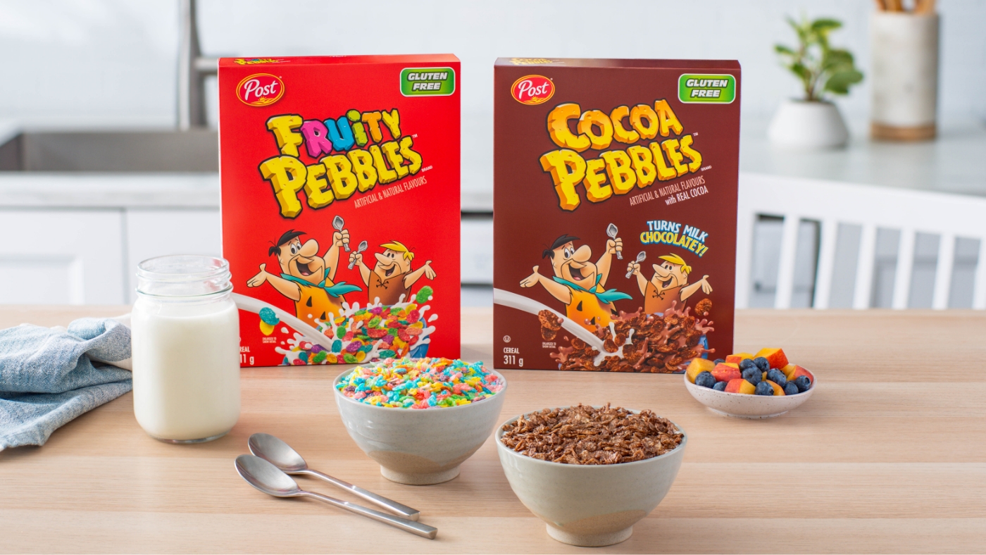Fruity Pebbles and Cocoa Pebbles cereal boxes sitting on a kitchen table with a jar of milk, two bowls of cereal and a plate of cut up fruit.