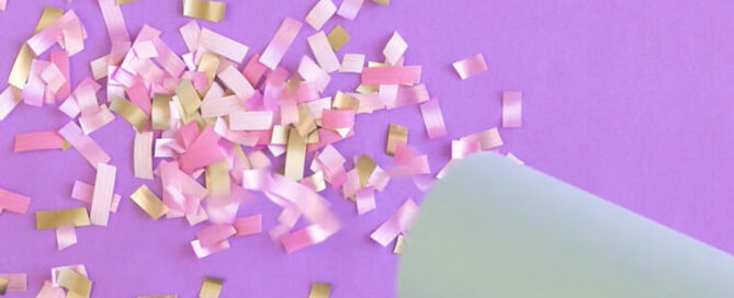Pink, white and yellow confetti on a purple background being blown from a cardboard tube.